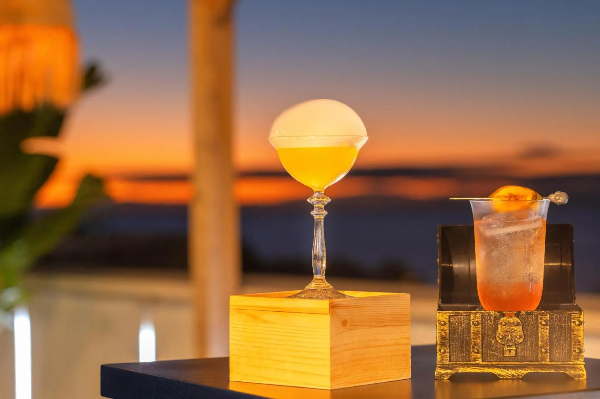 Un cocktail est présenté sur une table en bois avec une vue sur la mer au coucher du soleil.