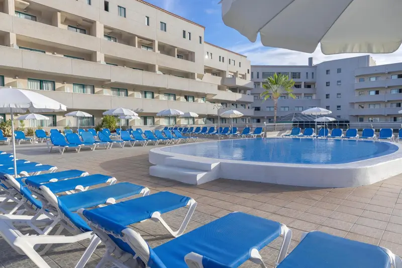 Transats bleus disposés autour d'une piscine ensoleillée devant un bâtiment blanc.