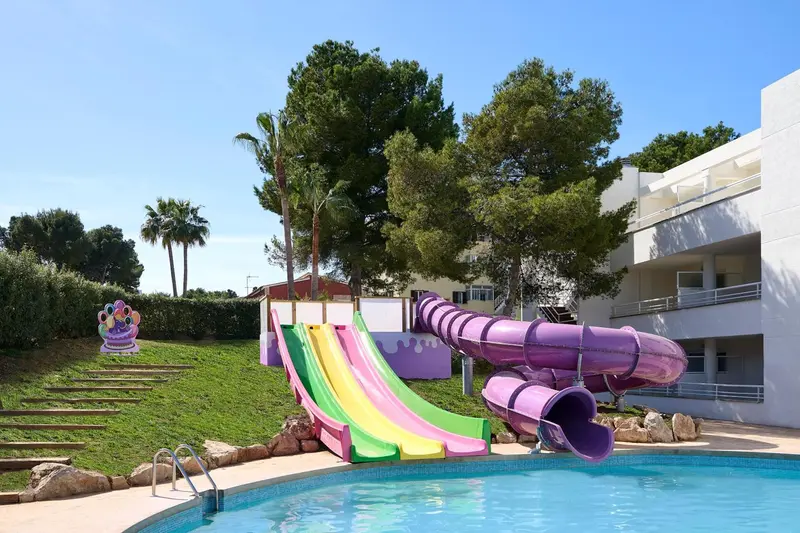 Toboggans colorés se terminant dans une piscine ensoleillée, avec une végétation luxuriante en arrière-plan.