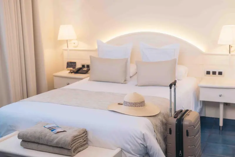 Une chambre lumineuse avec un grand lit et une valise posée sur le pied de lit.