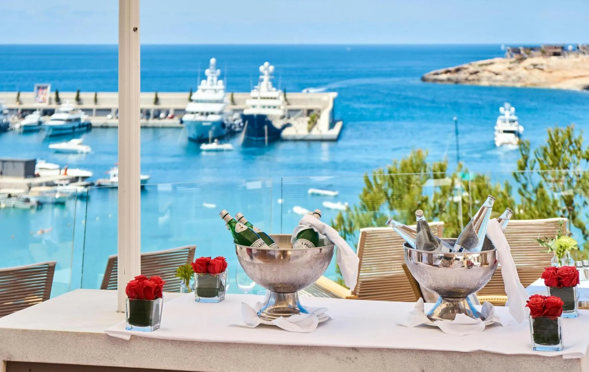 Une table blanche est mise en scène avec des bouteilles et des seaux à glace, face à un port de plaisance ensoleillé.