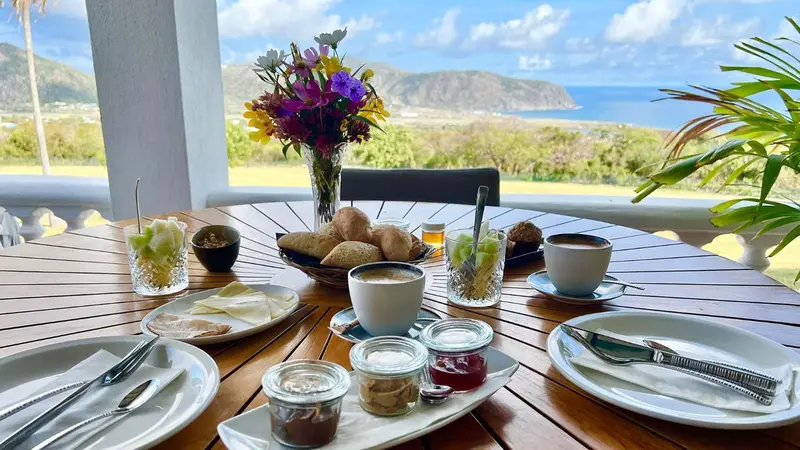 Une table en bois est dressée avec un petit-déjeuner face à un paysage montagneux et marin.