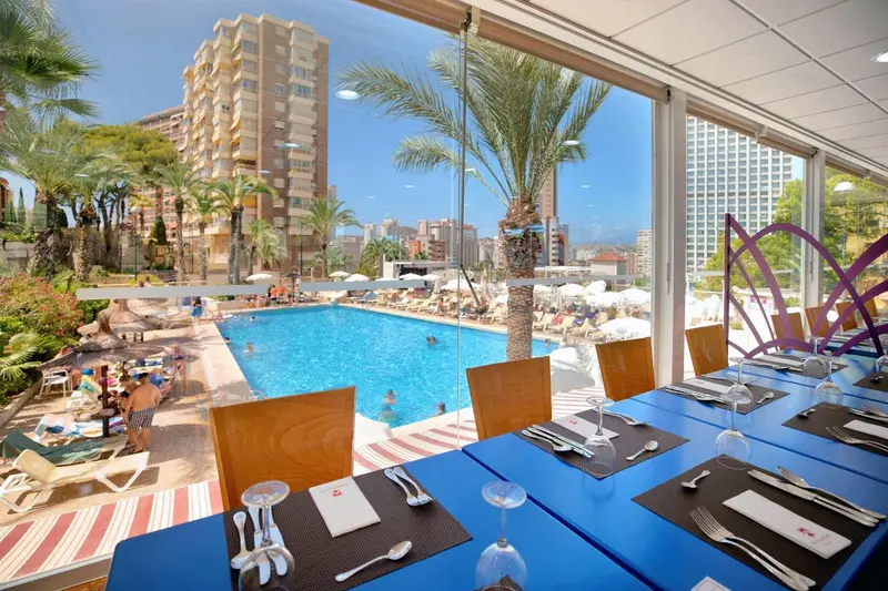Tables dressées en bleu face à une piscine ensoleillée et une vue sur la ville.