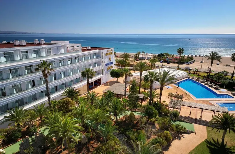 Vue aérienne d'un bâtiment blanc avec des balcons, une piscine et une plage ensoleillée.