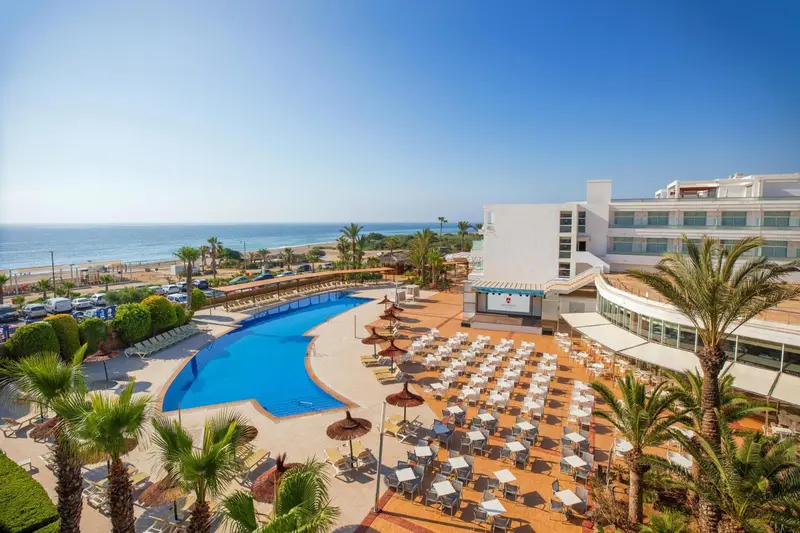 Vue aérienne d'une piscine d'hôtel avec des transats et des parasols, face à la mer.