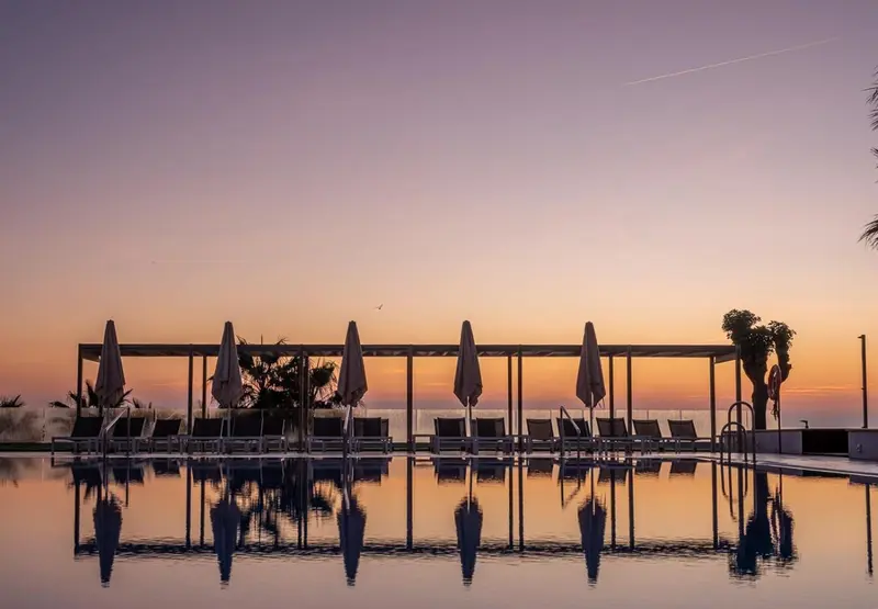 Transats et parasols alignés au bord d'une piscine reflétant le ciel crépusculaire.