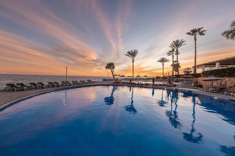 Une piscine bleue reflète le ciel orangé au coucher du soleil, bordée de chaises longues et de palmiers.