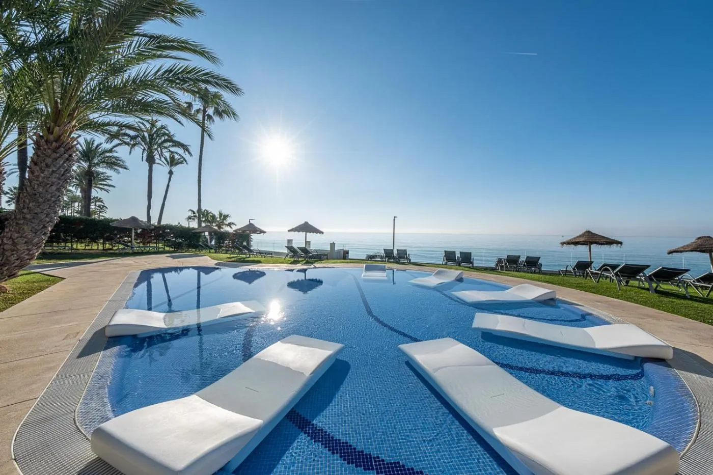 Transats blancs disposés autour d'une piscine bleue face à la mer.