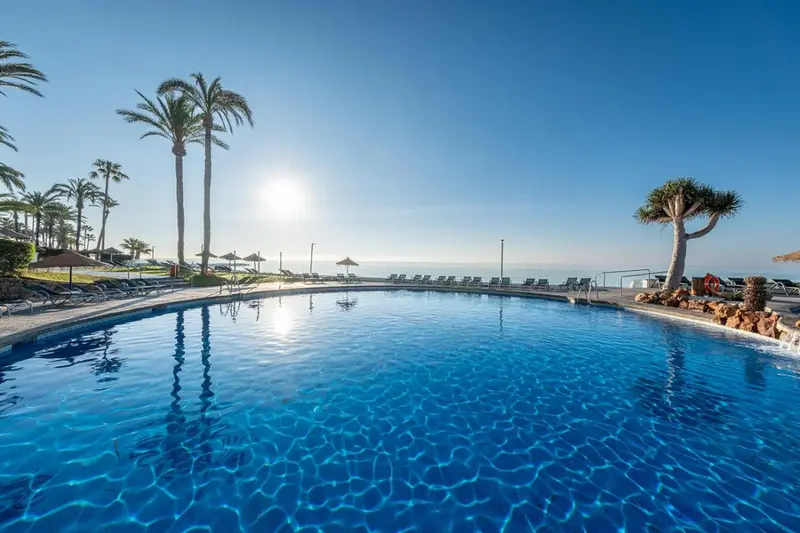Une piscine bleue bordée de transats sous un ciel ensoleillé.