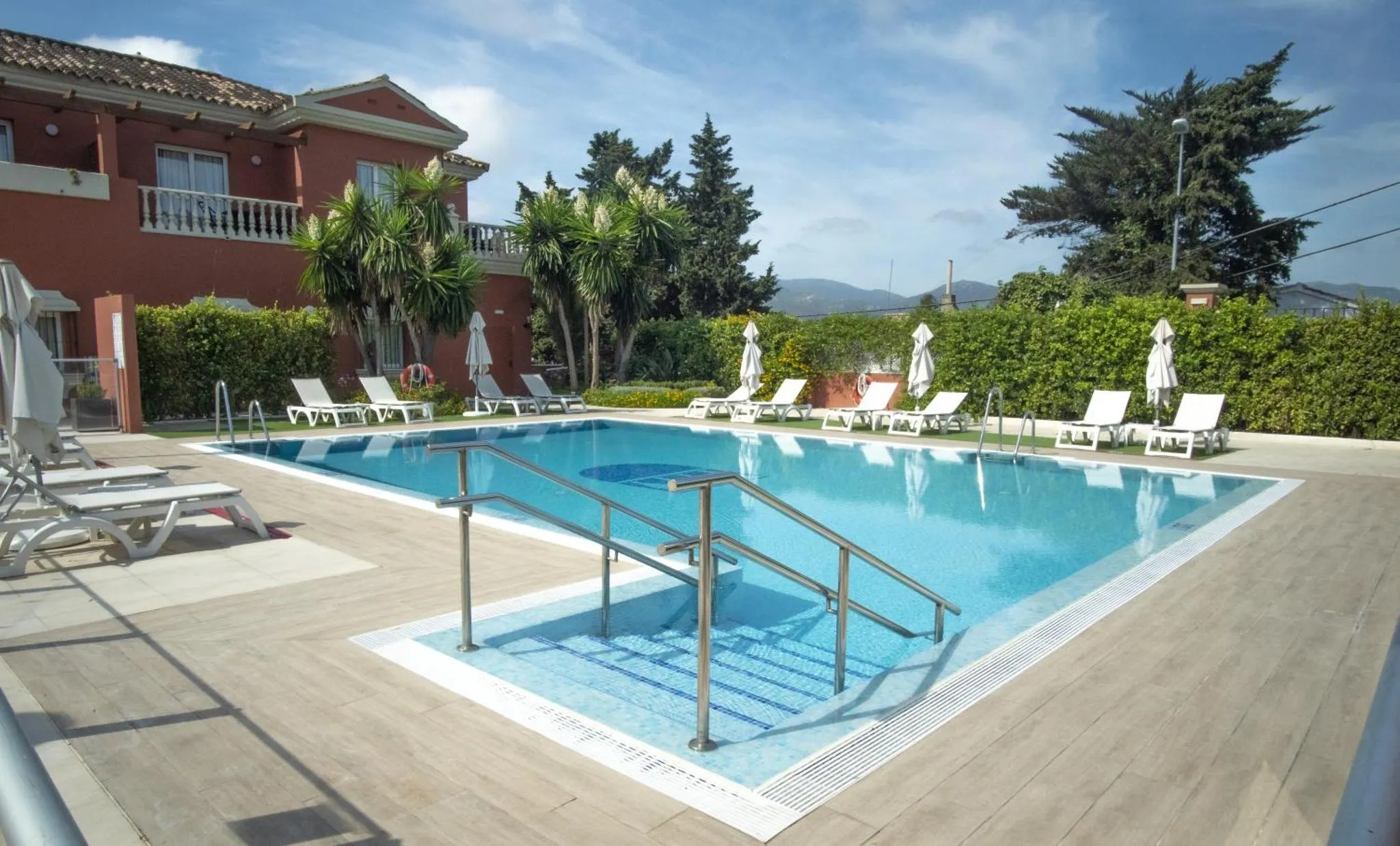 Une piscine rectangulaire bordée de transats et de palmiers, devant un bâtiment rouge.