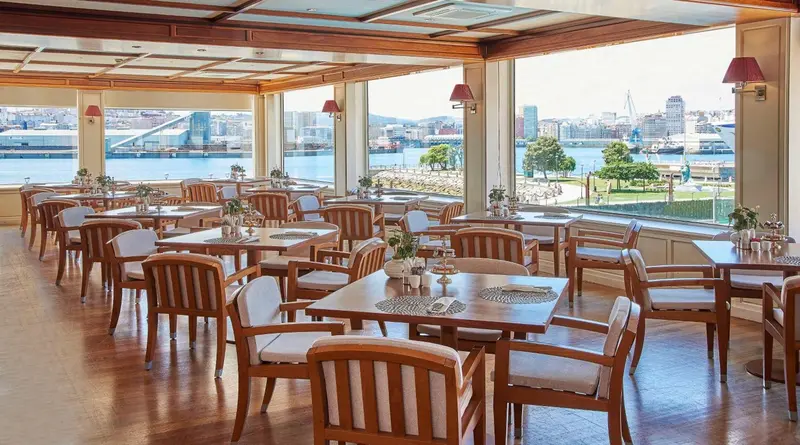 Une salle lumineuse avec des tables dressées face à une vue sur la mer et un port.