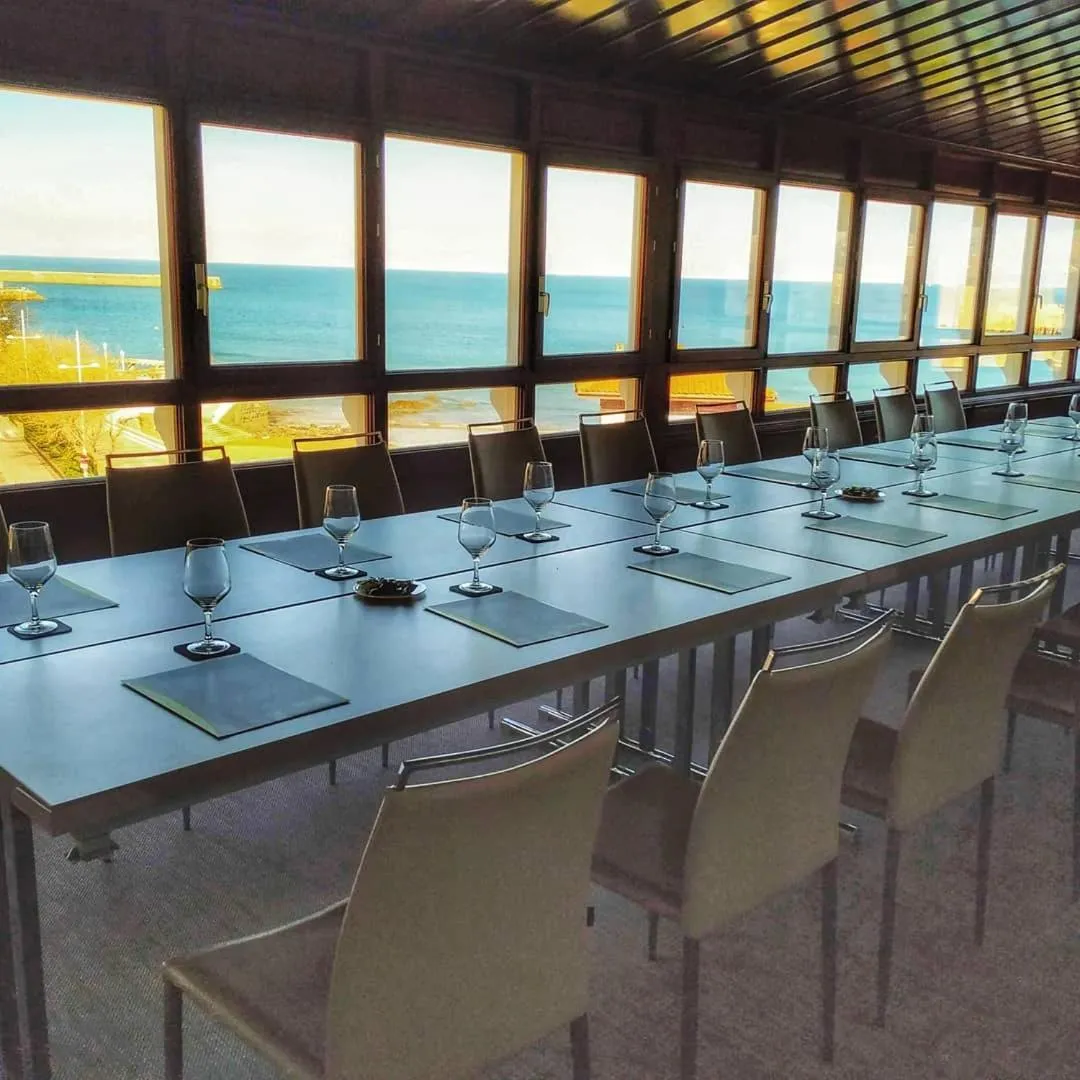 Une longue table de réunion est mise en place dans une salle lumineuse avec de grandes fenêtres donnant sur la mer.
