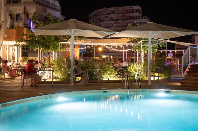 Terrasse lumineuse avec tables et chaises près d'une piscine éclairée.