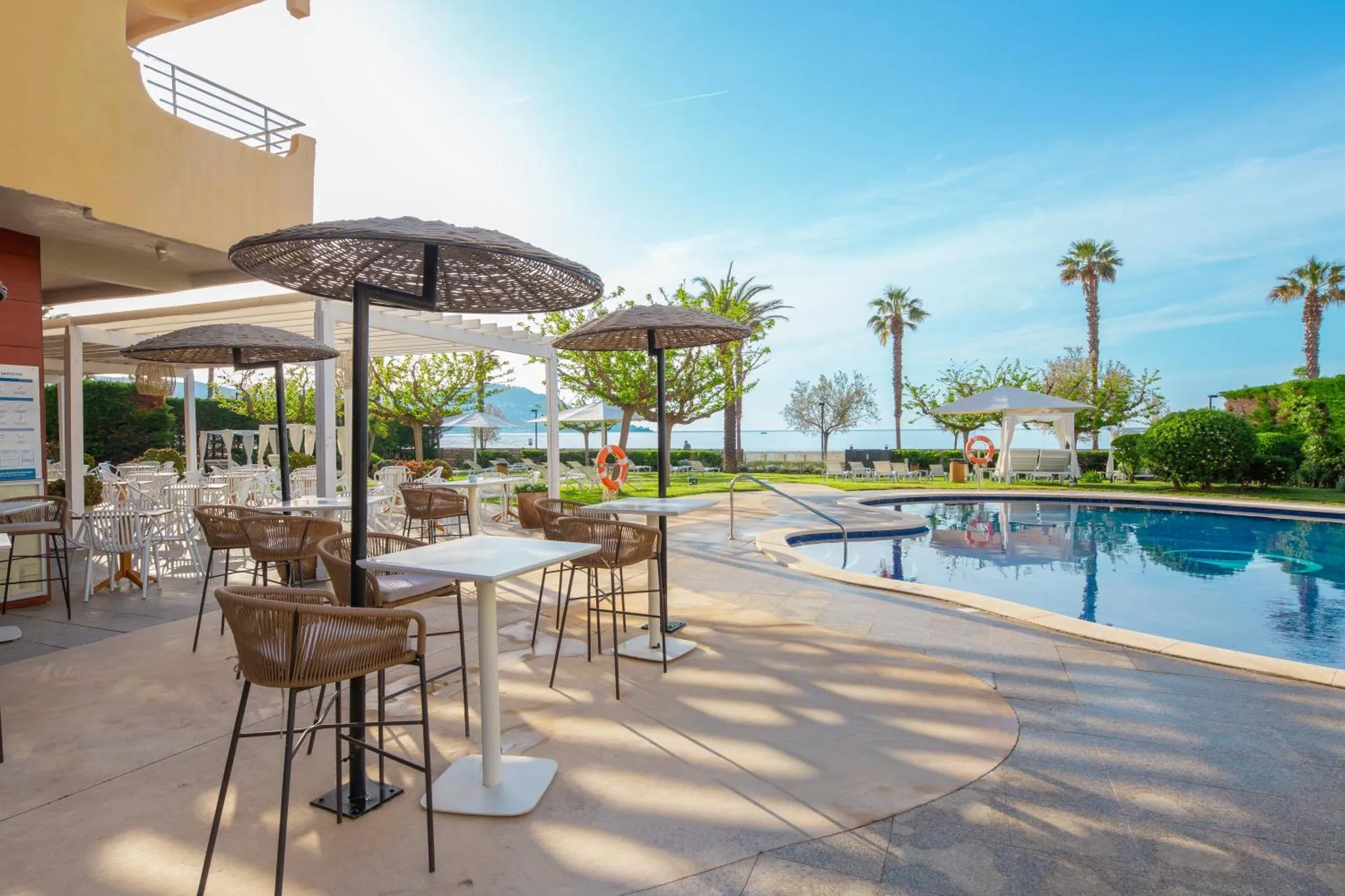 Terrasse ombragée avec tables et chaises près d'une piscine ensoleillée.