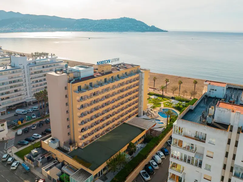 Vue aérienne d'un bâtiment hôtelier blanc avec de nombreux balcons, bordant une plage et une mer bleue.