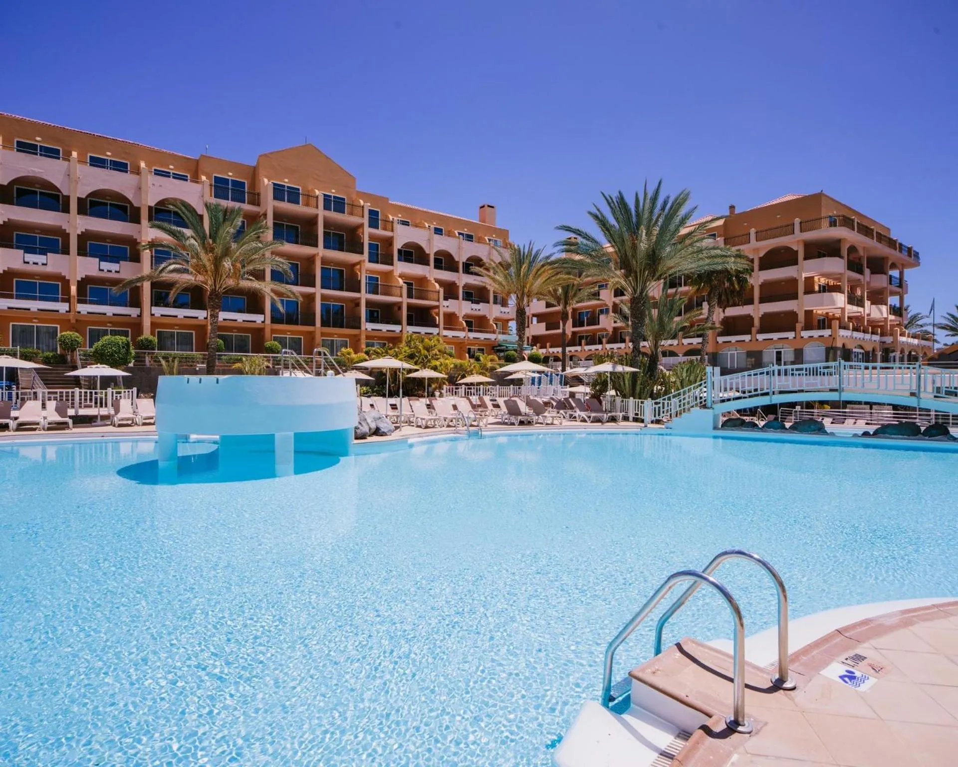 Piscine turquoise devant un bâtiment beige à plusieurs étages sous un ciel bleu.