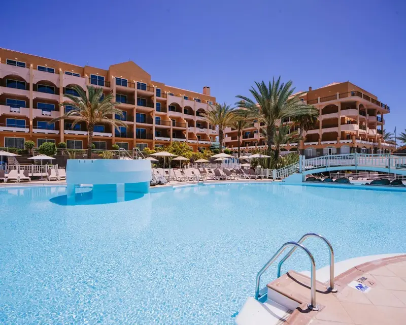 Piscine turquoise devant un bâtiment beige à plusieurs étages sous un ciel bleu.