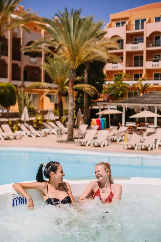 Deux femmes sourient et discutent dans un jacuzzi au bord d'une piscine ensoleillée.