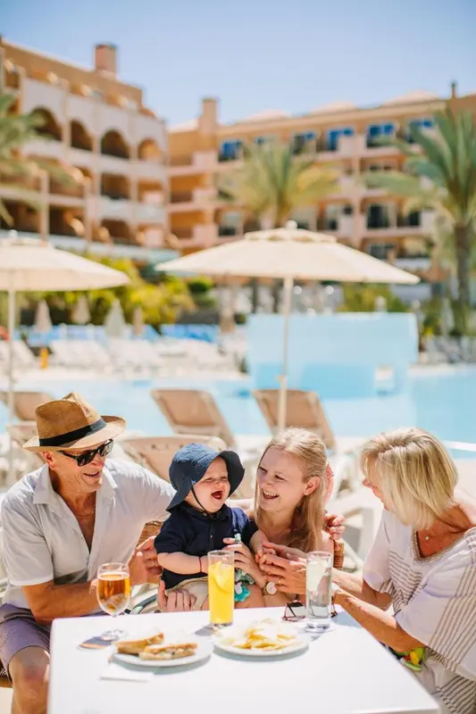 Une famille profite d'un repas en terrasse près d'une piscine ensoleillée.