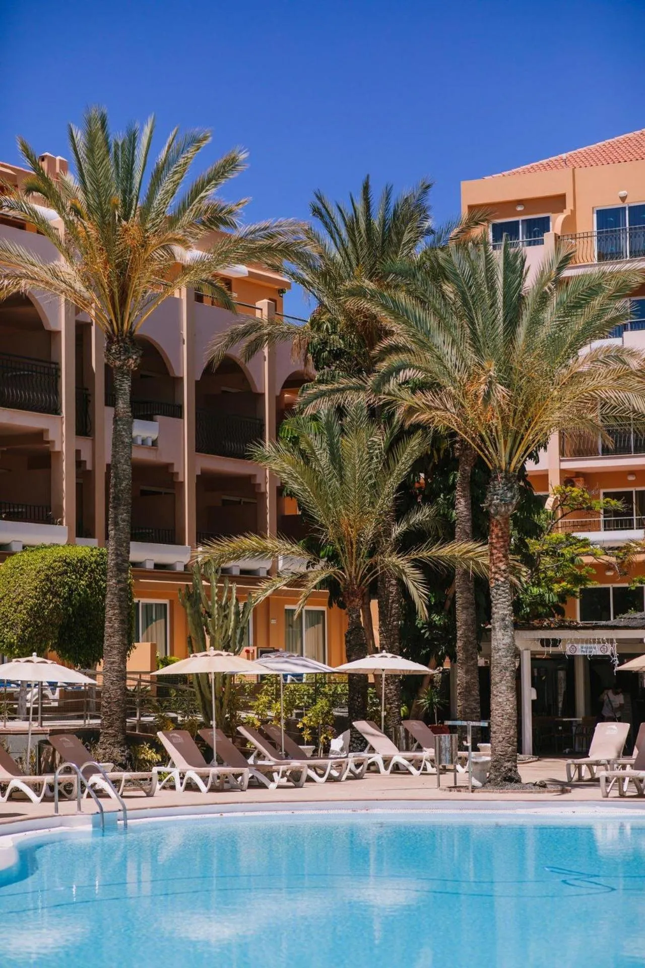 Transats et parasols bordent une piscine turquoise devant un bâtiment aux couleurs pastel et des palmiers.