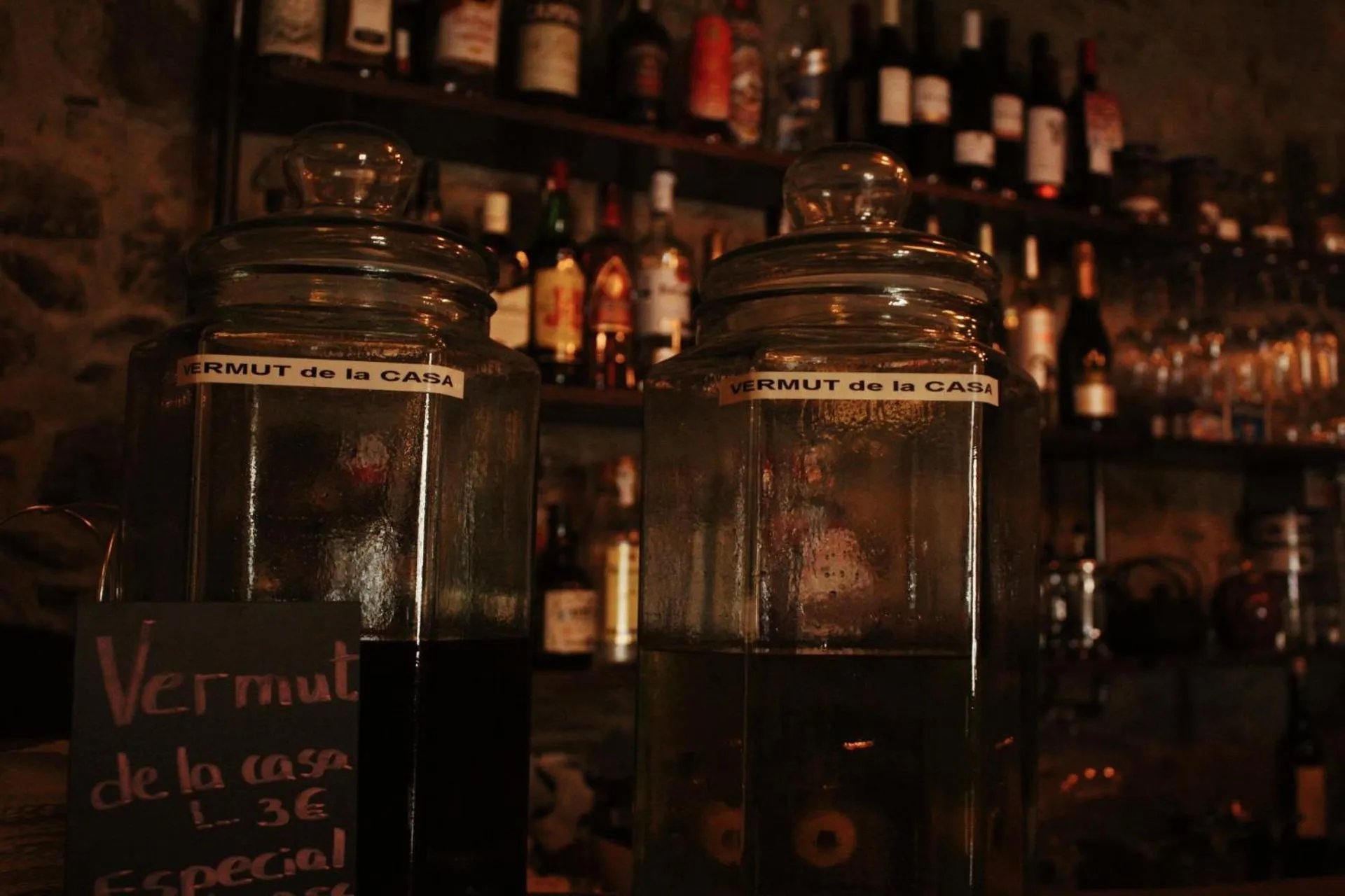 Deux grands bocaux en verre remplis de liquide sombre reposent sur un comptoir, devant une étagère garnie de bouteilles.