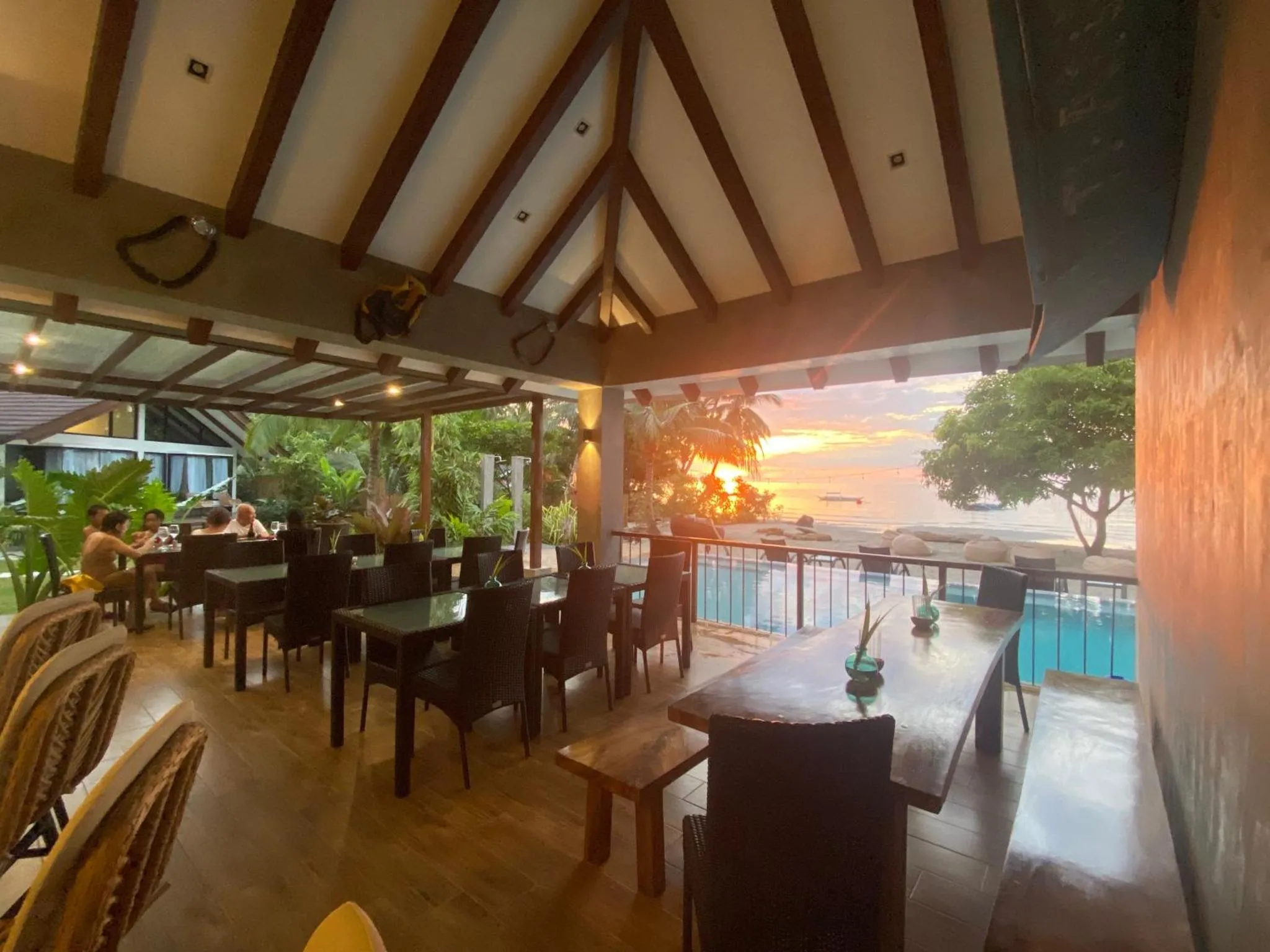 Terrasse ombragée avec tables et chaises donnant sur une piscine et la mer au coucher du soleil.