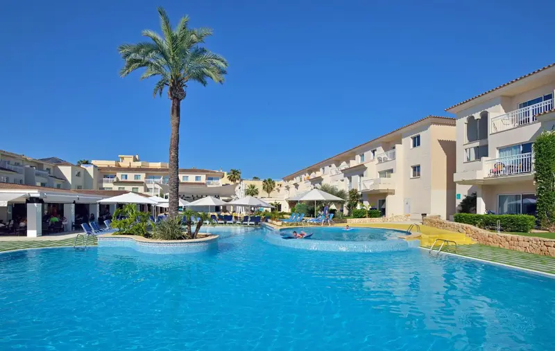 Vue d'une piscine d'hôtel entourée de bâtiments beiges et de palmiers sous un ciel bleu.