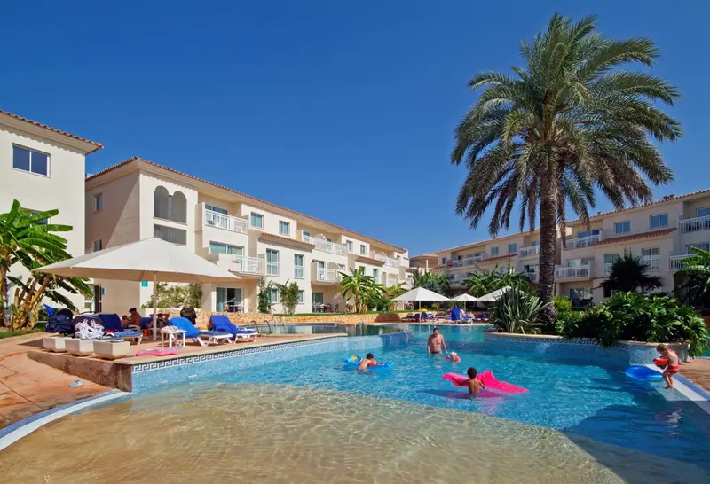 Une piscine bordée de transats et de palmiers devant un bâtiment beige.