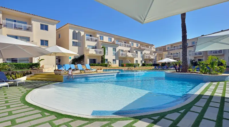 Piscine ensoleillée avec transats et bâtiments beiges en arrière-plan.