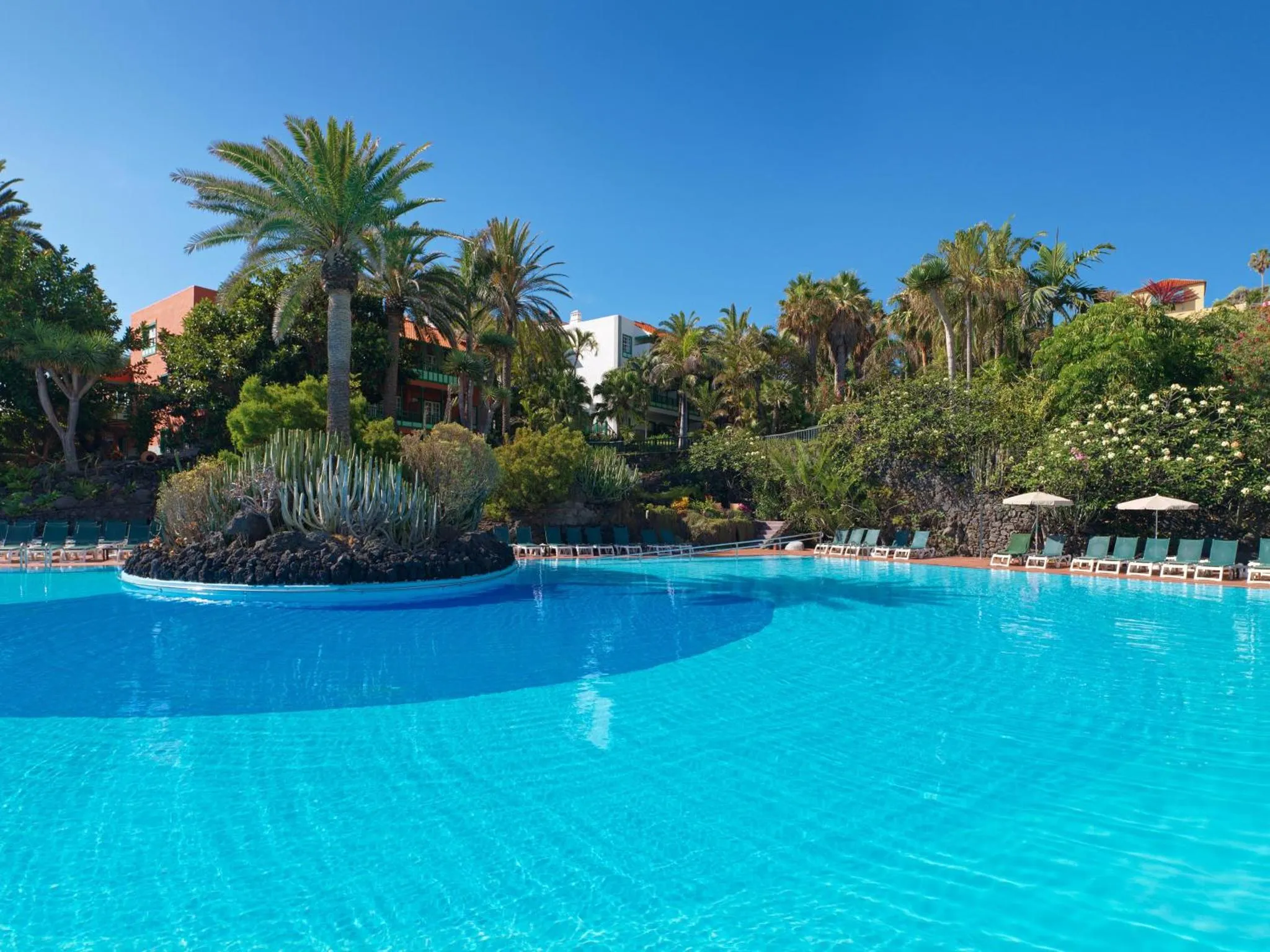 Une piscine turquoise bordée de transats et de palmiers sous un ciel bleu.