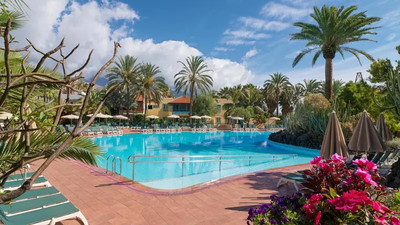 Une piscine rectangulaire bordée de transats et de végétation luxuriante sous un ciel bleu.
