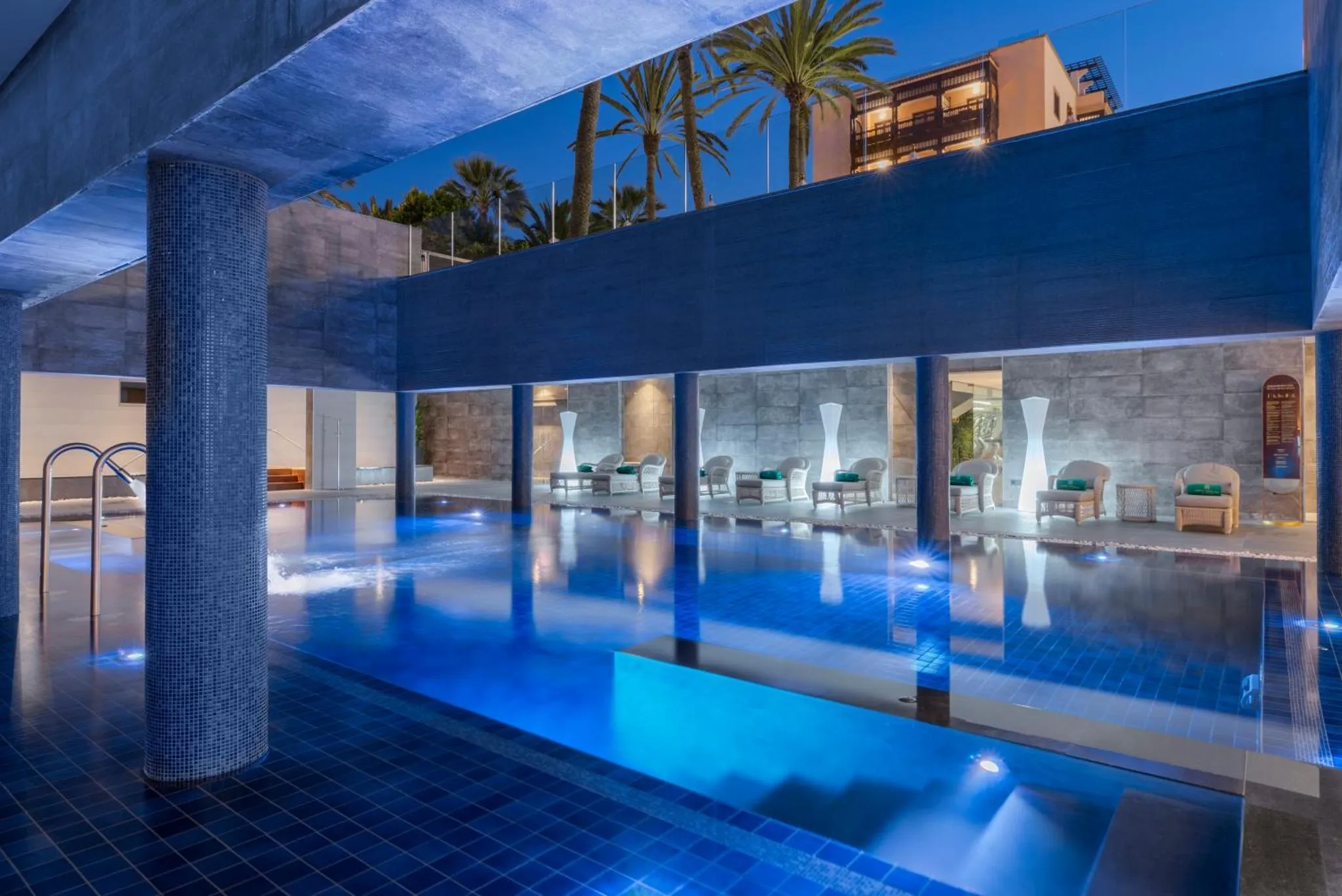 Piscine illuminée avec mobilier de détente et végétation tropicale en arrière-plan.