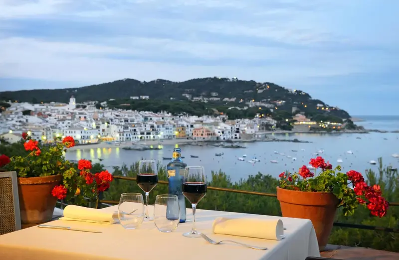 Table dressée avec des verres de vin et une vue sur une ville côtière illuminée au crépuscule.