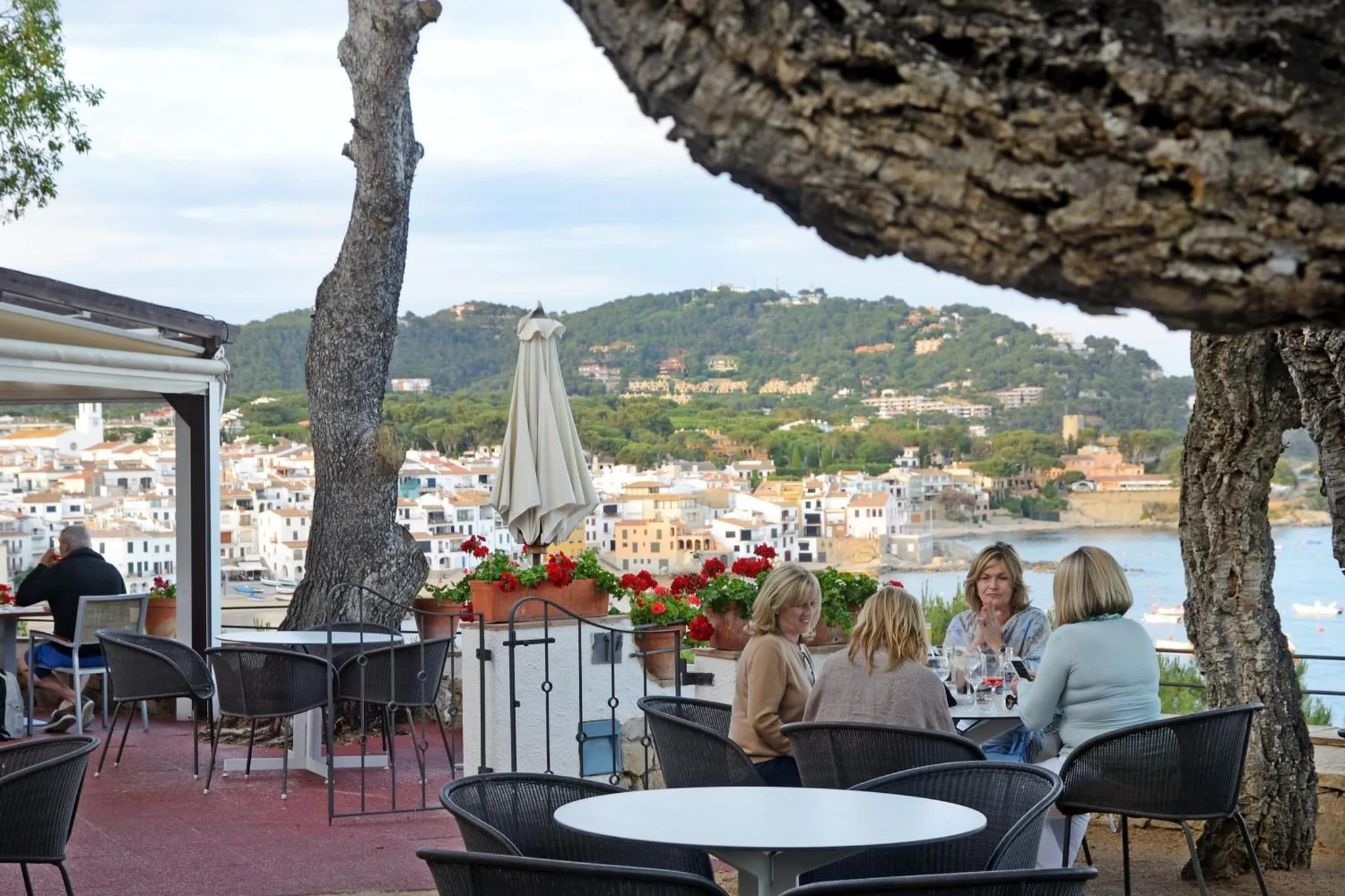 Des convives sont assis à des tables sur une terrasse ensoleillée avec vue sur la mer et la ville.