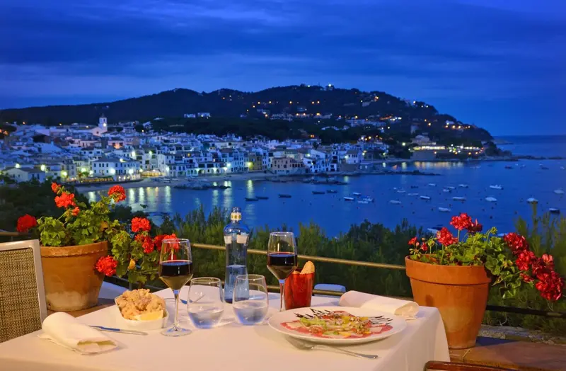 Table dressée avec des verres de vin et des assiettes en terrasse avec vue sur la ville côtière illuminée.
