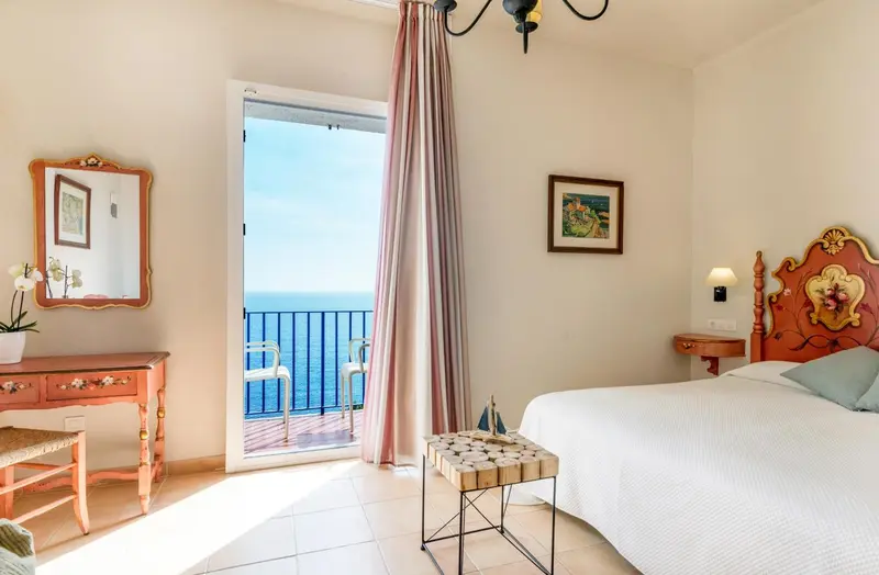 Chambre lumineuse avec un lit blanc et vue sur la mer par une porte-fenêtre.