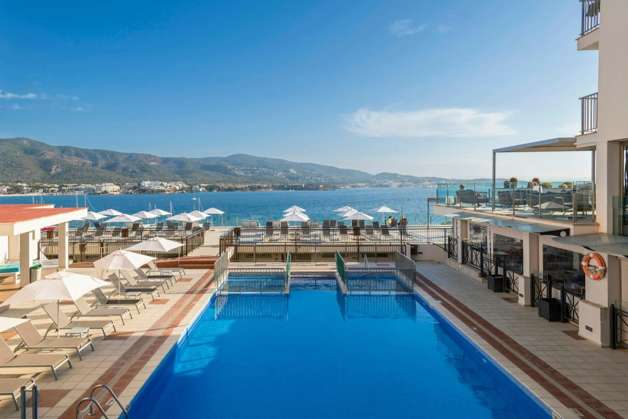 Une piscine bleue bordée de transats blancs offre une vue sur la mer et les montagnes.