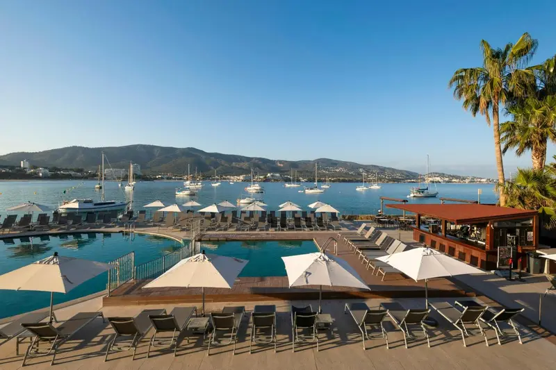 Transats et parasols bordent une piscine avec vue sur une baie ensoleillée.