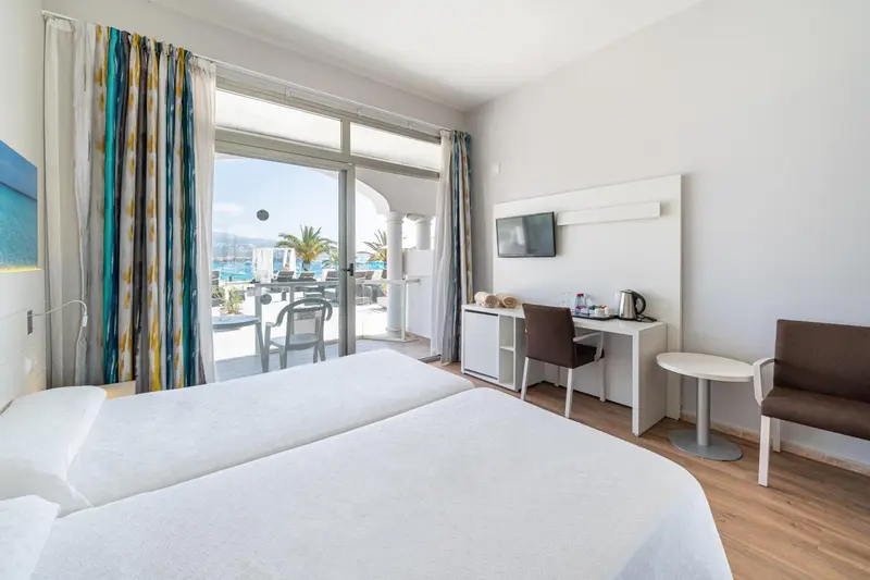 Chambre lumineuse avec un lit double et une vue sur une terrasse et la mer.