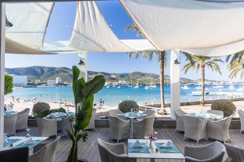 Terrasse de restaurant avec vue sur une baie ensoleillée et des montagnes.