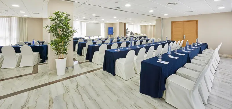 Une grande salle lumineuse est aménagée avec des tables et des chaises pour une réunion.