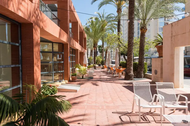 Terrasse ombragée avec tables et chaises devant un bâtiment aux façades colorées.