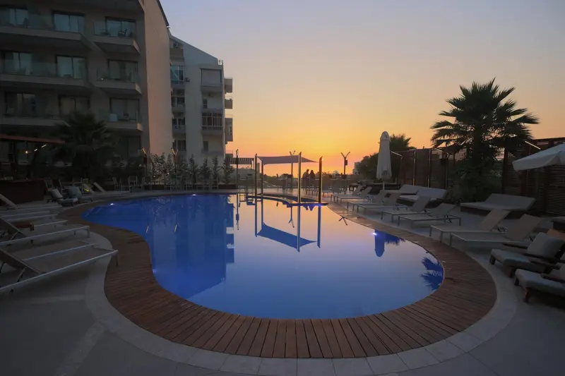 Une piscine bordée de chaises longues devant un bâtiment moderne au coucher du soleil.