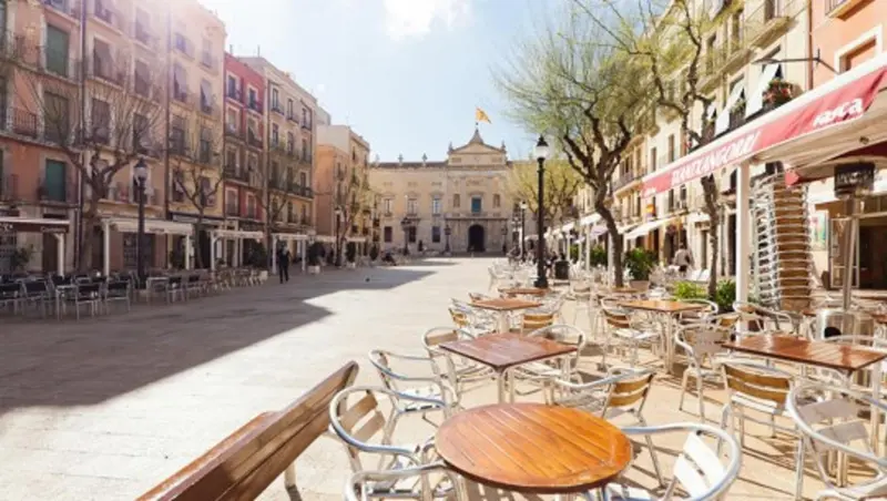 Une place ensoleillée avec des terrasses de café et des bâtiments aux façades colorées.