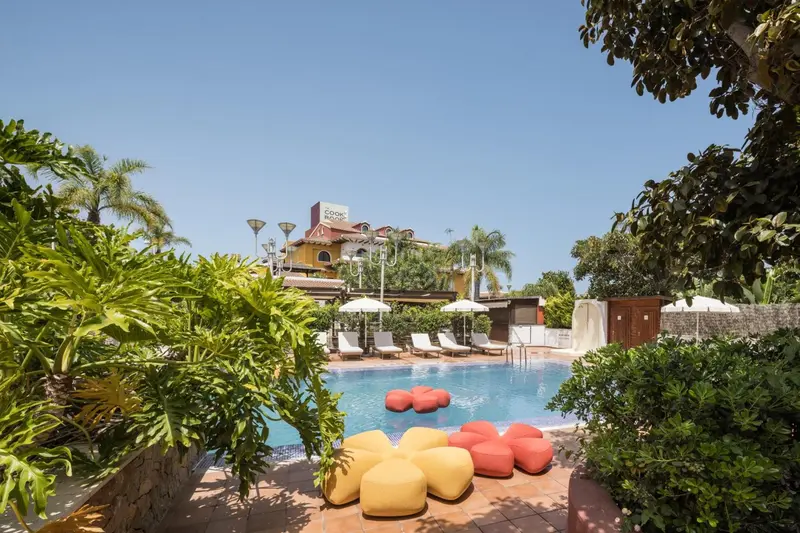 Une piscine bordée de chaises longues et de mobilier coloré, avec un bâtiment en arrière-plan.