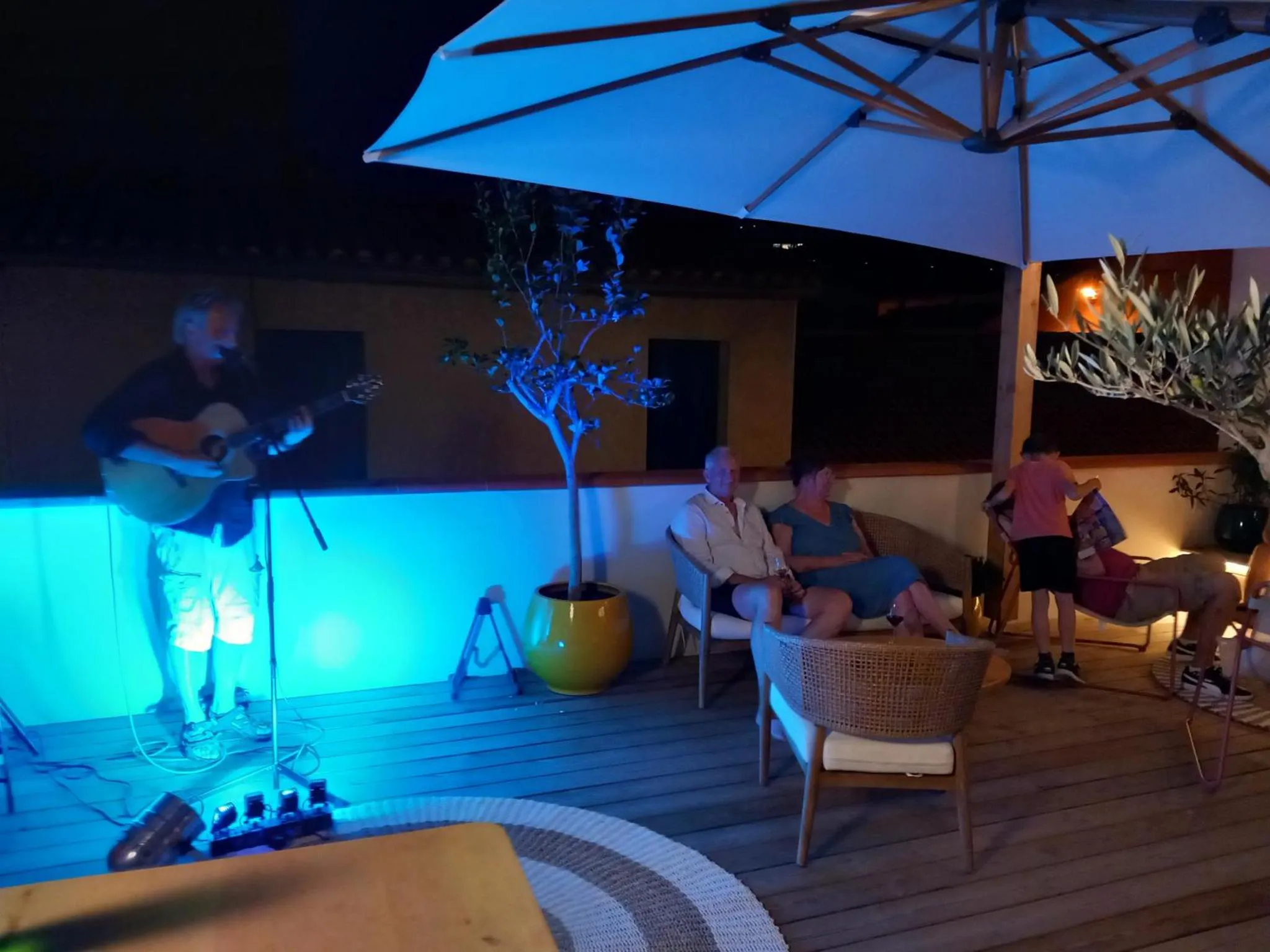 Un musicien joue de la guitare devant des convives assis sur une terrasse éclairée.