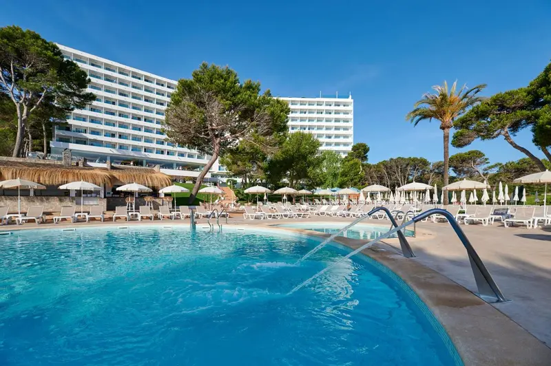 Transats et parasols bordent une piscine turquoise devant un bâtiment blanc.