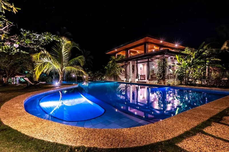 Une piscine illuminée reflète un bâtiment tropical la nuit.