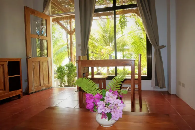Une table et des chaises sont disposées devant une grande fenêtre ouverte sur une végétation luxuriante.
