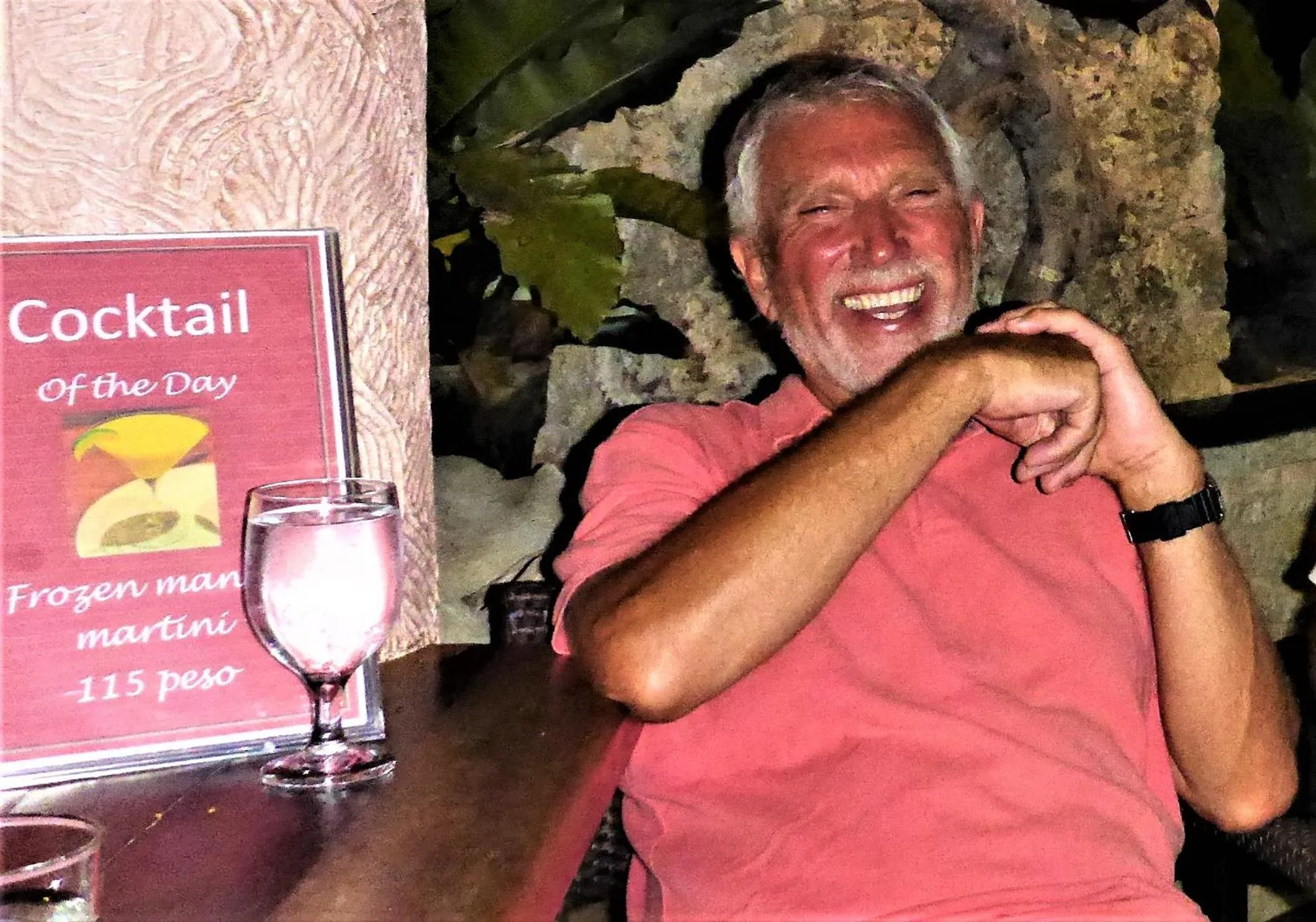 Un homme souriant est assis à une table avec un cocktail et un menu.